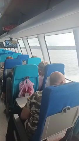 The video shows the interior of a passenger ferry. Rows of blue and teal seats are visible, some occupied by passengers. Luggage and bags are placed on seats and the floor. A pink plastic bag hangs from the back of a seat. A TV screen is mounted on the ceiling, displaying an image. Large windows offer a view of the water and land in the distance. The lighting suggests it is daytime.