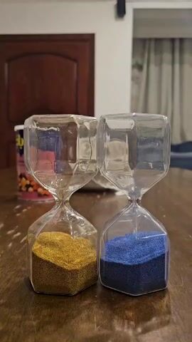 Two hourglasses are shown on a wooden table. One hourglass contains gold sand and the other contains blue sand. In the first few seconds, hands are holding the hourglasses and inverting them, causing the sand to fall. After the initial inversion, the sand continues to fall in both hourglasses. The scene appears to be indoors, possibly in a dining room or living room, with a wooden door and curtains visible in the background.