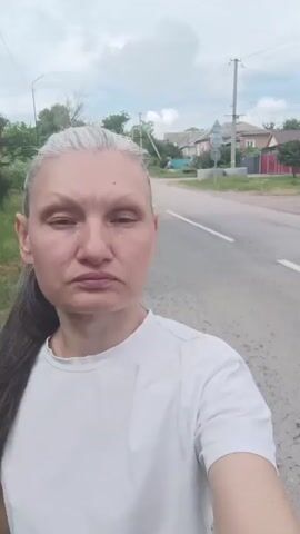A woman with graying hair pulled back in a ponytail and wearing a white t-shirt is walking on a paved road. She is looking directly at the camera with a serious expression. The road is empty except for a white line marking the lane. On the left side of the road, there is a dirt shoulder with dry grass and bushes. On the right side of the road, there are houses and trees. The sky is overcast.
