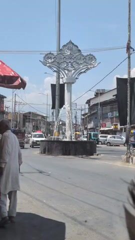 The video shows a busy street scene with a prominent fountain in the center. Cars and a three-wheeled vehicle are visible on the road, indicating traffic. Several buildings line the street, and utility poles and wires are present, suggesting an urban environment. The sky is clear and blue, indicating daytime. The fountain has a decorative structure at the top, possibly an artistic sculpture. A person is seen walking past the fountain. The overall impression is a vibrant street with typical urban activity.