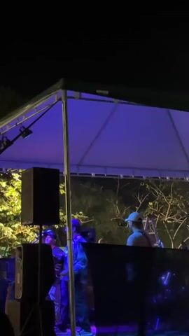 The video shows a band performing live outdoors at night, under a large tent illuminated with blue and pink lights. The lead singer, a man with a beard and a patterned shirt, stands at the microphone. A guitarist, wearing a cap, is positioned to the right of the singer. Speakers are located on the left, and other musical equipment is visible behind the stage. Trees are behind the stage.