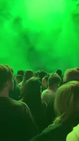 The video shows a large crowd of people at an event, possibly a concert, with bright green lights and fog filling the space. People are standing shoulder-to-shoulder, looking toward the stage. The camera moves through the crowd, focusing on different individuals, some of whom appear attentive and engaged. The background shows a packed stadium with many people in the upper tiers. The green lighting washes over everyone, giving the scene a surreal and energetic feel.