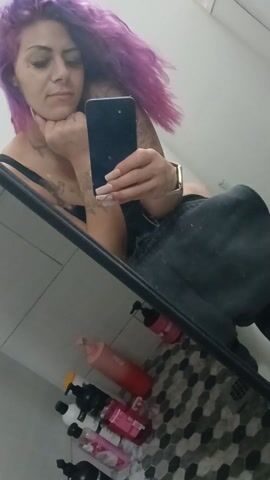 A woman with purple hair is taking a selfie in a bathroom mirror. She is wearing a black top and black pants. Her fingernails are long and painted. She has tattoos on her arms. The bathroom has a black and white tile floor. There are several bottles of shampoo and conditioner on the floor. She makes different expressions and gestures at the camera.