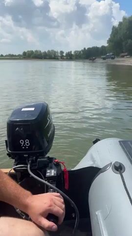The video shows a person's hand on a tiller connected to a black outboard motor with a "9.8" label. The motor is attached to the back of a white inflatable boat. The boat is on a body of water with a green hue. In the background, there is a shoreline lined with trees. A few vehicles, including what appears to be a truck and a yellow car, are parked on the shore. The sky is blue with scattered white clouds. The camera perspective is from inside the boat, looking out towards the shore and the sky. The boat is moving slowly through the water, creating small ripples.
