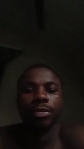 The video shows a close-up of a young Black man speaking directly to the camera in a dimly lit room. He is shirtless, and his face is the primary focus. He gestures with his left hand, pointing his index finger towards the camera. The background is dark, with some indistinct shapes visible that might be furniture or a wall. The man's expression appears earnest as he speaks, and the lighting suggests it is either nighttime or a very dark indoor setting.