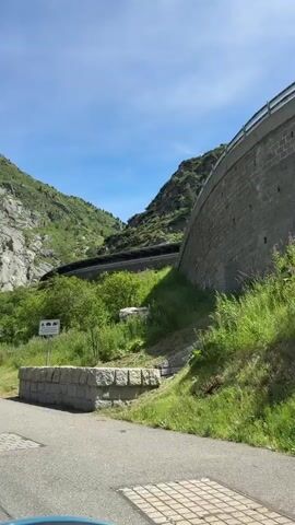 The video captures a scenic drive through a mountainous region on a clear, sunny day. The camera pans to the right, revealing a winding road carved into the side of a lush green mountain. A massive concrete dam wall, constructed with large stone blocks, dominates the right side of the frame, stretching upwards and disappearing out of view. Below the dam, dense green vegetation and tall grasses cover the slopes. In the middle ground, a section of the road is visible, covered by a long, dark structure that appears to be a tunnel or protective roofing, likely for the road. A white signpost with various symbols and text is placed in the grassy area on the left. The sky above is a clear blue with a few wispy white clouds. The overall impression is one of a well-maintained road passing through a dramatic natural landscape, possibly a mountain pass or a hydroelectric facility.