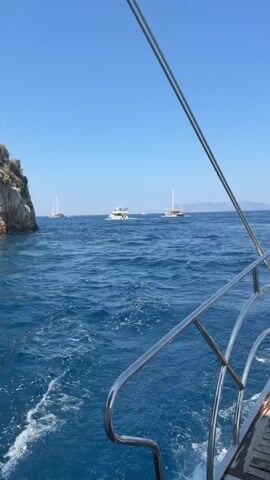 The video captures a view from the back of a boat moving through the ocean on a clear, sunny day. The water is a deep blue with whitecaps indicating movement. On the left side of the frame, a rocky, green-topped cliff juts out of the water. In the distance, several other boats are visible, including a sailboat and two smaller motorboats. The camera is positioned at an angle, looking out over the water, with the railing of the boat visible in the foreground. The sky is a vibrant blue, with no clouds. The boat is leaving a white wake behind it.