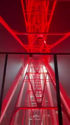 The video shows the inside of a Ferris wheel car, looking out at the metal structure of the ride. The Ferris wheel structure is illuminated with red lights. A ladder is visible in the center of the structure. The background appears foggy. Someone is speaking in the background, but their words are unclear.