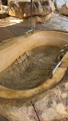 The video shows a stone-like tub filled with muddy water. Metal fixtures extend into the tub. The tub is situated in an outdoor area with other stone structures and foliage. A small waterfall can be seen in the background. The camera zooms out to reveal more of the surrounding area, including a shaded structure over the tub. There are tools and other items scattered around the base of the tub.