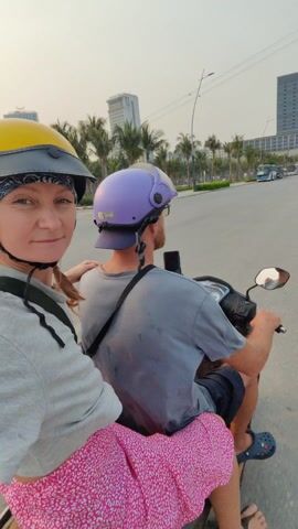 A woman is filming from the back of a scooter driving down a street in what appears to be a Vietnamese city. The woman is wearing a yellow helmet, a grey t-shirt, and a pink skirt with a pattern. The driver, a man with a purple helmet and a grey t-shirt, is navigating the vehicle. The street is lined with palm trees and modern buildings. Other vehicles, including scooters and buses, can be seen on the road. The sky is overcast. The woman looks at the camera and smiles at one point during the ride.