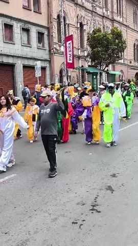 A group of people wearing colorful clown outfits are walking down a street in a parade. They are wearing bright colors like red, green, yellow, purple, white, and blue, and many are wearing hats and sunglasses. Some of them are carrying yellow batons. Behind the parade, there are people walking and watching. The buildings on either side of the street are multi-story and made of stone, with arched windows. There is a red banner with the word "PACEÑA" hanging from one of the buildings. The sky is overcast.