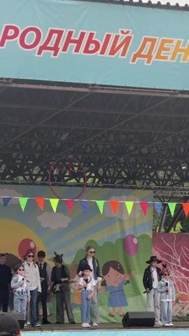 Here's a description of the video:

The video shows a group of children and young adults on a stage. A banner above reads "Mezhdunarodnyy Den," which translates to "International Day." The backdrop is decorated with a colorful mural featuring a sun, clouds, and cartoon children. A string of colorful triangular pennants hangs across the front of the stage roof.

The group on stage consists of about ten people, mostly dressed in casual outfits. Some of them are wearing sunglasses. At least one person is holding a guitar. Some members have animal ear headbands on. The stage itself appears to be an outdoor structure with a metal framework visible above. The audience is partially visible in the foreground, with the backs of heads and shoulders appearing at the bottom of the frame.