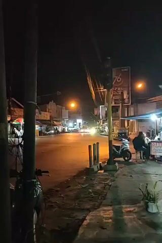 The video shows a street scene at night, illuminated by streetlights and the lights from shops. A traffic light is visible, cycling through its signals. Several motorbikes are parked on the side of the road, and two people are sitting at a small table outside a shop. Cars are driving on the road in the distance. The overall atmosphere is one of a typical urban street at night.
