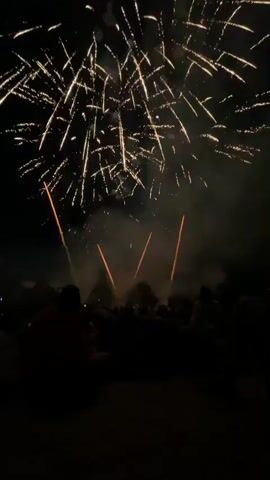 This media file captures a fireworks display at night in Canet de Mar, Spain. The scene is set on what appears to be a beach or sandy area, with a crowd of people silhouetted in the foreground, facing away from the camera and towards the sky. The people are seated or kneeling, suggesting a relaxed atmosphere as they watch the event unfold.

The main activity is the spectacular eruption of fireworks. Multiple bursts of golden and white light illuminate the dark night sky, creating intricate patterns of sparks that trail downwards. Wisps of smoke drift through the air, partially obscuring some of the lower bursts and adding to the dramatic visual effect. Occasional trails of orange light descend from some of the explosions, hinting at the trajectory of the pyrotechnics. The overall mood conveyed is one of awe and enjoyment, as is typical for fireworks displays. The time of day is clearly night, indicated by the blackness of the sky and the illuminated fireworks. There are no visible text elements in the media file. The location cues, such as the sandy ground and the clear night sky, are consistent with an outdoor event, and the provided context specifies Canet de Mar, Spain.