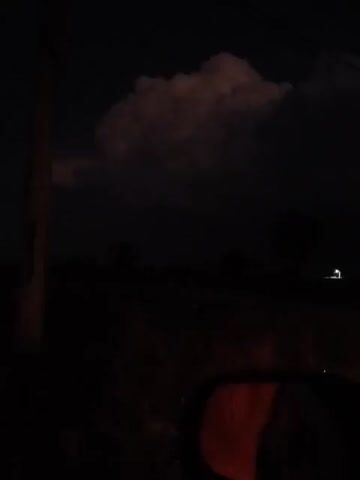 The video is taken at night from inside a car. A dark, cloudy sky is visible through the windshield. In the distance, a faint light can be seen. A utility pole is visible on the left side of the frame. The car's side mirror is visible in the lower right corner, reflecting a reddish light.