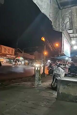 This is a low-light video of a street scene at night. A traffic light is visible, showing a red light. Cars are driving on the wet street. A scooter is parked on the right side of the frame. There are some food stalls on the right, and people are visible near the stalls. A building with lights is visible on the left side of the street. The overall atmosphere is that of a busy urban environment at night.