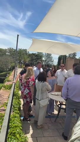 The video shows a group of people gathered outdoors on a sunny day, likely at a social event. They are standing on a patio or balcony with a brick railing covered in green vines. A white canopy provides shade. The people are dressed in a variety of casual and semi-formal attire. They are engaged in conversation and appear to be enjoying themselves. In the background, there is a well-manicured garden with trees and pathways. Some people are walking in the garden. A table with some food items is present on the patio.