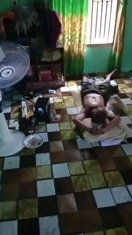 An overhead shot shows a man lying on his back on a checkered floor. He is on a pillow and appears to be resting. To the left of the man, a white electric fan is visible, along with a black speaker. A window with green curtains is in the background. The room appears to be a residential setting.