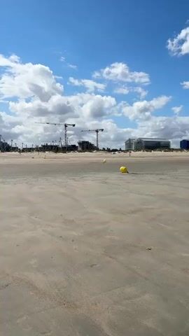 The video captures a wide, sandy beach under a bright blue sky with scattered white clouds. In the distance, there's a shoreline with calm, green water. The sand is textured with rippled patterns, suggesting the tide has recently receded. A few yellow buoys are visible on the sand, spaced out across the frame. In the background, along the distant horizon, there are signs of human activity: several construction cranes are prominent, indicating ongoing development. Beyond the cranes, some buildings are visible, including what appears to be a modern, glass-fronted structure and a large blue building. The scene is peaceful, with only the subtle presence of human construction to break the natural beach environment.