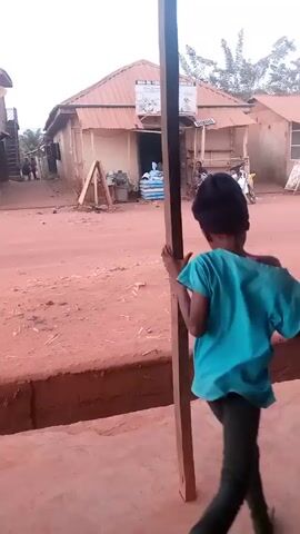 A young child playfully runs toward and holds onto a wooden support beam.  The child, dressed in a blue shirt, leggings, and a black head covering, is barefoot. The child playfully peeks around the beam at the camera and then runs away, only to quickly return, looking back and smiling.  The scene takes place on a dirt road in front of a building with a sign on it. In the background, there is a motorbike, other buildings, and some people walking.