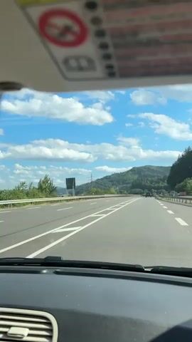 The video shows a view from inside a car driving on a highway. The road has multiple lanes and is bordered by a guardrail and a grassy embankment. Hills covered in trees are visible in the distance. The sky is blue with fluffy white clouds. The sun is shining, creating some lens flare in parts of the footage. There are a few other cars on the road, and some buildings and infrastructure can be seen along the side. The interior of the car, including the dashboard, windshield, and side mirror, are also visible.