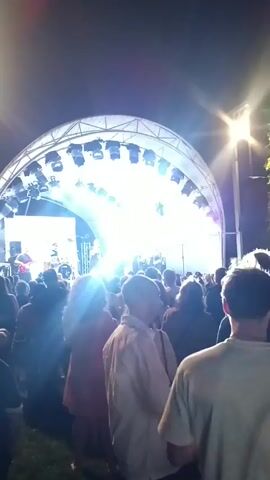 A crowd of people are gathered in front of a stage at night.  Bright lights illuminate the stage where a band is performing. The crowd appears to be enjoying the performance.