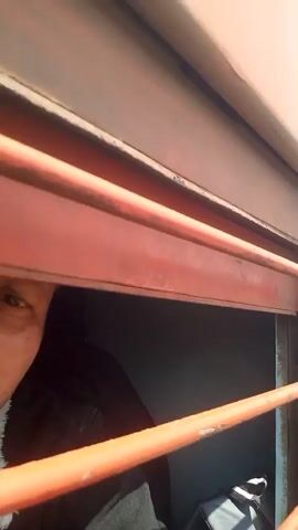 The video shows a person looking out of a train window, which then pulls back to show the exterior of the train moving along the tracks. Other trains are visible on parallel tracks, and overhead power lines and support structures are also present. The environment around the tracks appears to be a railway yard with scattered debris.