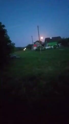 The video is of a rural landscape at dusk or night. The sky is dark blue, transitioning to black. A bright, pale moon is visible in the sky, partially obscured by a telephone pole. In the middle ground, there are several houses with lights on. A tree is visible on the left side of the frame. The foreground is dark, with blurred grass. There is no discernible movement or activity in the video.