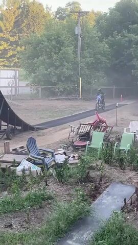 A person on a blue dirt bike jumps off a ramp in a rural setting. The bike is in the air and the rider is wearing a helmet and protective gear. The background shows trees and a clear sky. The person lands on a large inflatable airbag.