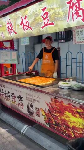 This media file shows a street food vendor in Linghe, China. The vendor, wearing an orange apron and a red cap, is cooking a large rectangular dish on a flat-top grill. The dish appears to be a type of tofu or similar food item, cut into squares and arranged in a grid. The vendor is using a spatula to tend to the food.

The stall is decorated with red and yellow signage featuring Chinese characters. One sign above the vendor's head reads "香豆," which translates to "fragrant bean," and other characters likely refer to the food being sold. The stall itself is brightly lit, and the surrounding area suggests a bustling night market or commercial street.

In the background, other food stalls and vendors are visible, along with a crowd of people walking through the area. The lighting indicates it is likely evening or dusk. The overall atmosphere appears lively and active, typical of a vibrant street food scene. Signs and decorations feature red lanterns and imagery related to traditional Chinese culture. The food stall also displays images of various skewers and fried food items.