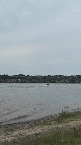 A wide shot captures a body of water with a distant shoreline. On the water, a jet ski is in motion, leaving a wake behind it. The shoreline is lined with houses and trees, suggesting a town or village. The sky above is overcast, with soft, diffused light. The foreground shows a rough, grassy embankment. The overall scene is tranquil and appears to be a summer day.
