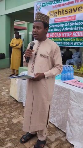 A full-shot video captures a man standing in front of a banner and addressing an audience. He is wearing a light brown kaftan and a patterned cap, holding a microphone and a document. The banner, partially visible, has text indicating "KADUNA STATE MINISTRY OF HEALTH" and "Sightsavers," along with a message about drug administration and promoting safe water. Behind him, another man in a yellow garment stands with his arms crossed. Several other people are seated in the background. The setting appears to be outdoors, with a paved ground and greenery visible. The man is speaking and gesturing, suggesting he is giving a speech or presentation.