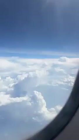 The view from an airplane window shows a dense layer of white, puffy clouds beneath a bright blue sky. The curvature of the airplane window is visible in the bottom right corner of the frame. The sun is shining above, indicating daytime. The scene is static with no visible movement other than slight variations in the clouds.