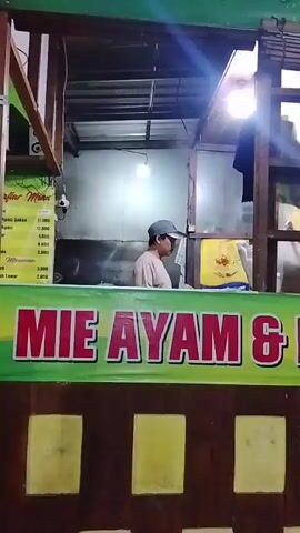 A person wearing a light-colored t-shirt and a grey baseball cap is standing behind a food stall. The stall has a green banner with red text that reads "MIE AYAM & BAKSO". There are yellow squares on the front of the stall. A motorcycle is parked to the right of the stall. The stall is lit by an overhead light.