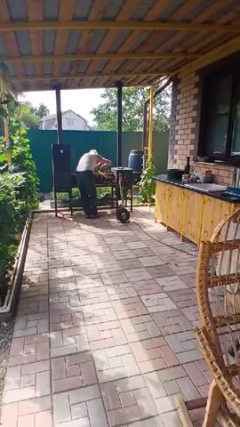 A person is standing in front of a grill. They are wearing a light-colored tank top and dark pants. The grill is black and has a metal grate. There are skewers of food on the grill. The person is holding a tool, possibly for turning the food. The grill is on a paved patio. There is a covered area above the patio, supported by yellow metal poles. To the left of the grill is a green fence and some plants. To the right is a brick building with a wooden counter and some bottles on it. The sun is shining.