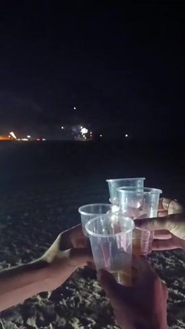 A nighttime beach scene with four people holding up plastic cups for a toast. In the background, fireworks are exploding in the sky over the ocean. The sand is visible in the foreground.