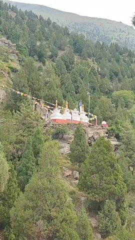 A drone flies over a mountainous landscape covered in trees. Several Buddhist stupas and buildings are visible on a hillside. Prayer flags are strung between the structures.