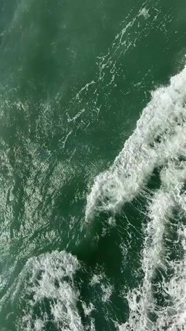 The video is an aerial view of the ocean. The water is a deep green color with white foam from waves breaking on the right side of the frame. The waves appear to be moving from the bottom right of the frame towards the top left. The texture of the water is rough and choppy.
