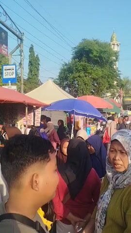 A bustling outdoor market scene unfolds under a clear blue sky. Tents, some with vibrant red and blue umbrellas, are set up, displaying various goods. A diverse crowd of people, many wearing hijabs and head coverings, navigates the crowded aisles. In the background, a minaret of a mosque rises above lush green trees, and a building with Islamic architectural elements is visible. The atmosphere appears lively, with individuals interacting and browsing the market stalls.