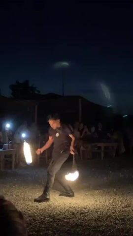 A young man is performing a fire juggling act at night. He is wearing dark clothing, a necklace, and boots. He is holding two burning torches, one in each hand, and is in the process of spinning them. Flames are visible from both torches. He is standing on a gravel-covered ground. In the background, there are tables and chairs set up, and several people are seated, some holding up phones, likely recording the performance. There are also some structures with lights in the background. The sky is dark blue, indicating it is nighttime.