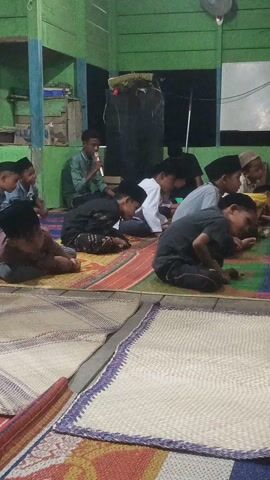 The video shows a group of people, mostly young boys, sitting on mats on the floor of a room. The room appears to be simple, with green wooden walls and a few objects visible in the background, including a fan and some shelves. The people are dressed in casual clothing, and some of the boys are wearing traditional Islamic hats. They appear to be engaged in some kind of religious activity, possibly a prayer or a lesson, as they are looking towards the front of the room, where a person is standing and speaking. The lighting is dim, suggesting it might be evening or nighttime. The overall atmosphere seems to be one of reverence and learning.