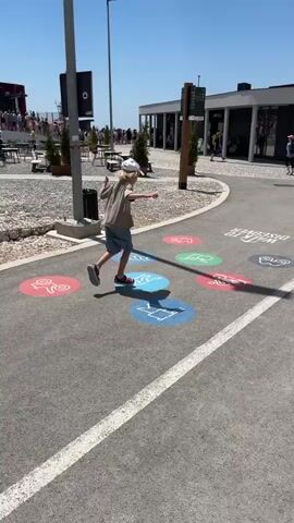 The video shows a young child wearing a hat, t-shirt, and shorts, playing a game of hopscotch on a paved area outdoors on a sunny day. The hopscotch court has colorful circular markings with various icons, including what appears to be a sailboat, a bicycle, a person running, and other symbols. The child is in the process of hopping from one circle to another, holding a frisbee. In the background, there are buildings with shops, tables and chairs outside, trees, and other people walking around. The environment appears to be a public recreational area or a promenade.
