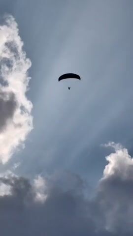 A paraglider is shown in silhouette against a partly cloudy sky. The paraglider is small in the frame and appears to be descending. The clouds are scattered, with some areas of bright light and some areas of shadow. The overall impression is one of open space and freedom.
