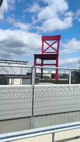 The video shows a large red chair sculpture in an outdoor setting. The camera appears to be moving, possibly from a vehicle, as indicated by the blurred foreground elements. The chair is the dominant feature, situated against a backdrop of a blue sky with white clouds. In the middle ground, there are buildings, trees, and a parking lot. Yellow banners with German text are visible on poles in front of the buildings. The overall scene suggests an urban or commercial area.