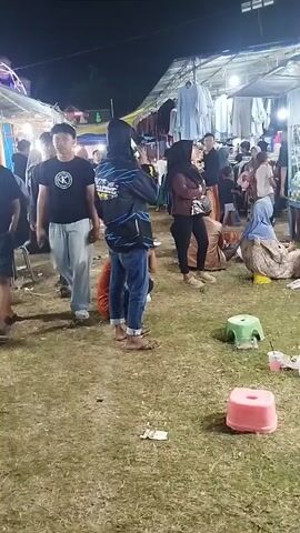 This video pans across an outdoor night market. In the foreground, a young man wearing a black t-shirt with a white logo walks towards the left. He is followed by another person wearing a black jacket with a blue design and a hood. There are several small plastic stools scattered on the grassy ground. To the right, a ball pit attraction is visible with many colorful balls. Several people are gathered around the ball pit and stalls. A table in the foreground is covered with various colorful toys. The market appears to be set up under temporary shelters with bright lighting.