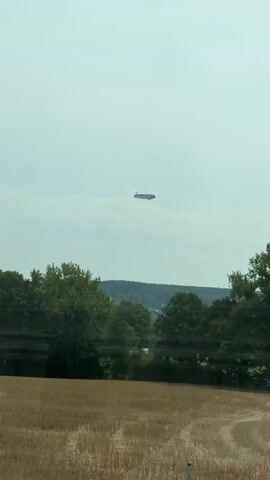 The video pans across a landscape with trees in the foreground and a hazy sky in the distance. An elongated, dark object is visible in the sky. The object appears to be either an aircraft or a blimp. The scene is likely outdoors during the daytime.