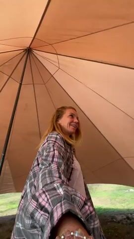 A woman with blonde hair is smiling and looking to the right inside a large, light brown tent. She is wearing a plaid shirt with pink and gray squares over a white shirt. The tent has a conical shape with radial seams. Outside the tent, there is grass and trees, and in the distance, there are other tents, one green and one light blue. The camera is positioned low and looking up.