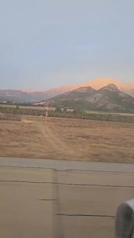 The video shows a view from an airplane window as it taxis on a runway. In the foreground, a portion of the airplane's orange and silver engine is visible. The runway extends into the middle ground, where dry, brown grass and a fence can be seen. In the background, a range of mountains is illuminated by the soft light of what appears to be sunrise or sunset. A windsock is also visible on the runway. The sky is a clear, pale blue. The camera is moving, indicating the plane is in motion.