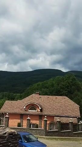 The video shows a blue car parked in front of a house with a tiled roof. The house is surrounded by a stone fence with decorative posts. In the background, there are lush green trees and hills under a cloudy sky. The scene appears to be a residential area situated in a natural, hilly landscape.