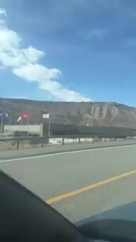 The video shows a view from inside a car moving on a highway. The sky is blue with some white clouds. On the right side of the road, there is a gray, almost barren hill or mountain range in the distance. Along the road there is a gray concrete barrier. Two flags are visible.